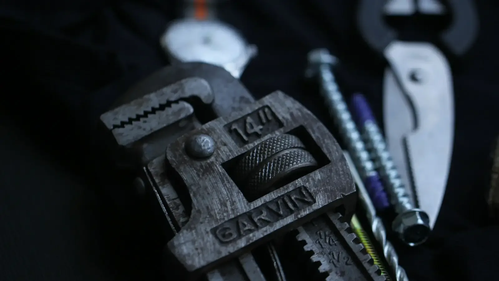 A worn pipe wrench among screws and pliers on a dark bench